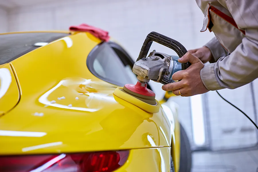 A technician using a power buffer polisher to repair and polish the rear quarter panel of a bright yellow sports car inside an auto body shop.